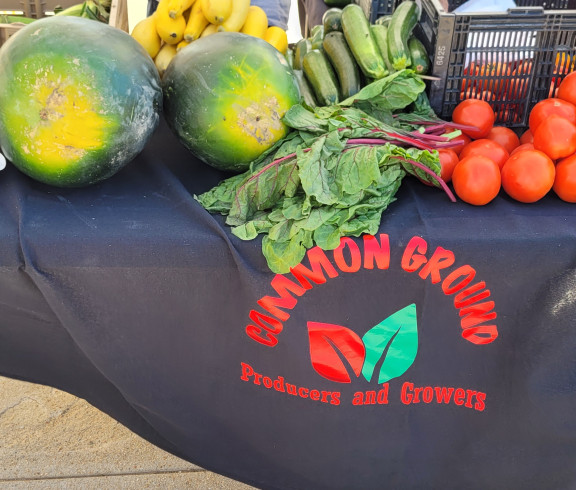 A table with a Common Ground Producers & Growers table cloth on it and full of produce (green beans, corn, summer squash, watermelon, zuchini, tomatoes). 